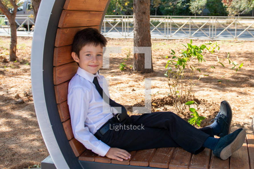a sweet little boy sitting in a park in dress clothes 