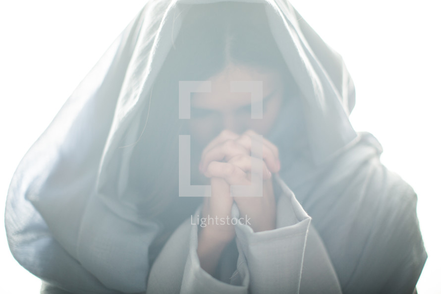 Mary in a blue shroud in bright light with head bowed and praying hands 