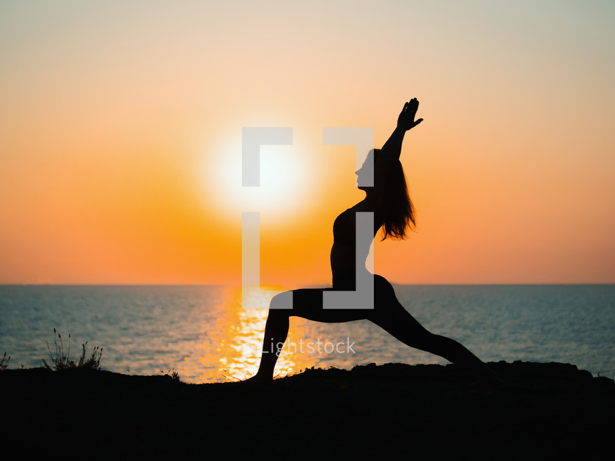 Young slim girl practicing yoga on mountain against ocean or sea at sunrise time. Silhouette of woman in rays of awesome sunset.