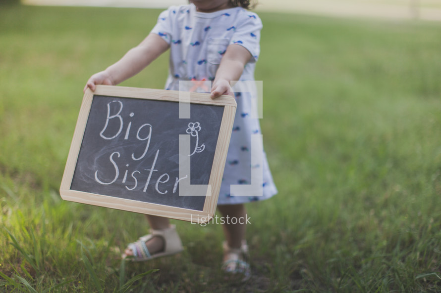 Girl holding a "big sister" chalkboard sign outside.
