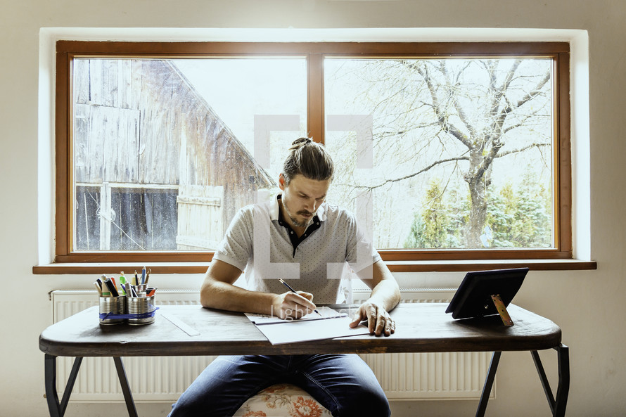 a man sitting at a desk drawing with a farm scene behind him 