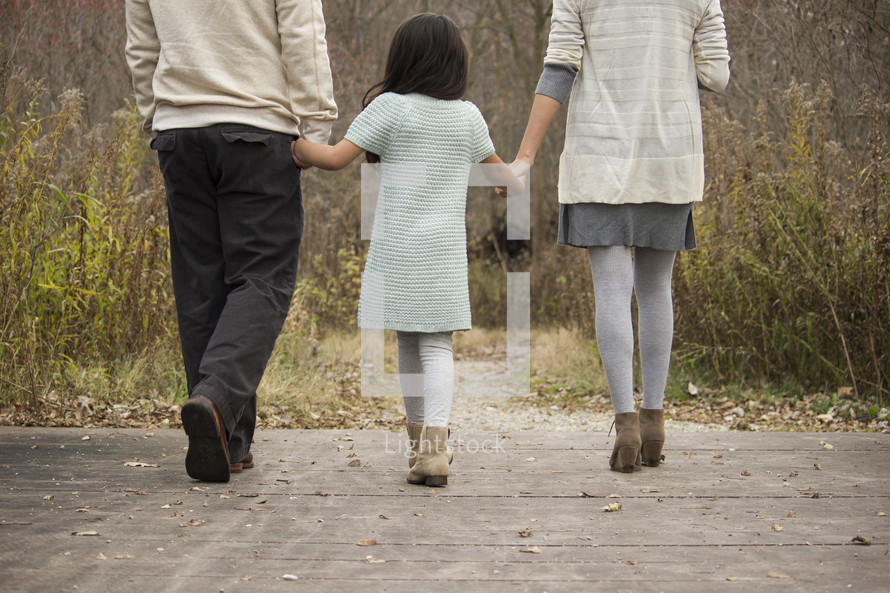 Family holding hands, walking across a wooden bridge outside.