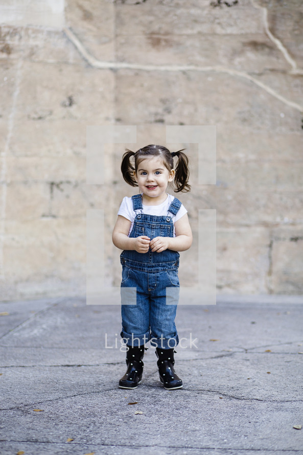 A little girl in overalls in front of a stone wall.