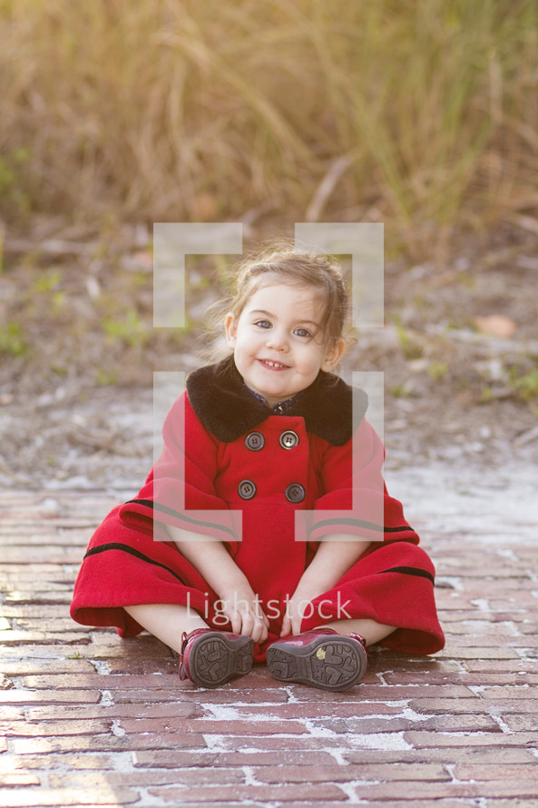 A young child in a red coat sitting on a brick path.