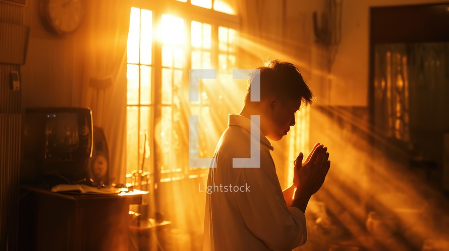 A man in a white shirt is praying in front of the window in the rays of light