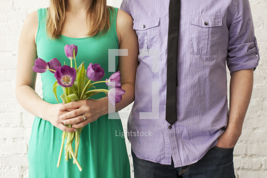 Couple standing against a brick wall, with the woman holding a bouquet of tulips.