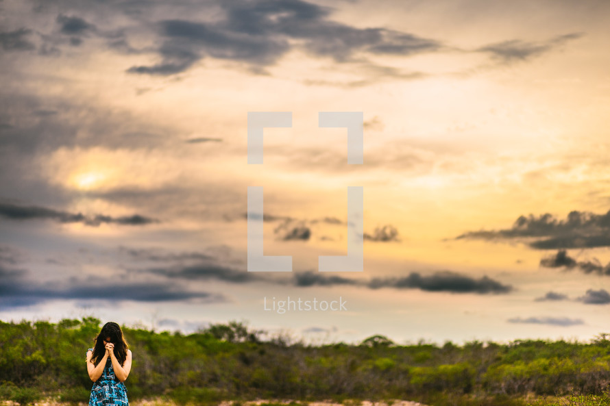 a woman praying with head bowed outdoors at sunset 