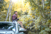 a young man sitting on top of his vehicle praying outdoors in a forest 