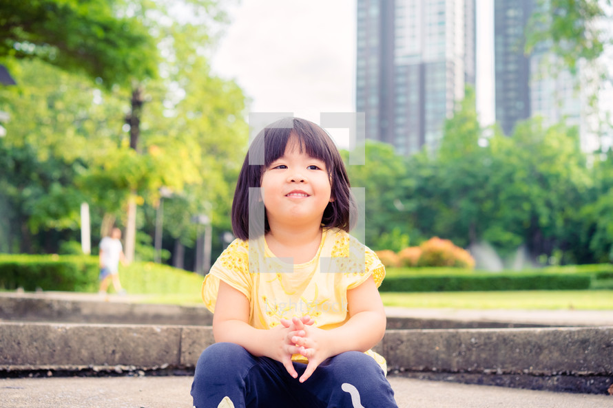 a toddler girl sitting in a park in a city 