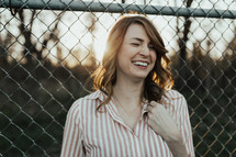 A laughing woman standing in front of a chain link fence.