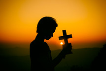 Young man praying and holding Cross at sunset  