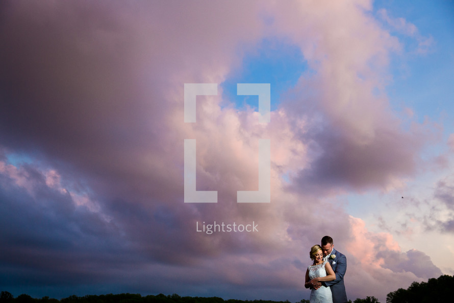 A bride and groom embrace outdoors under a cloudy sky.