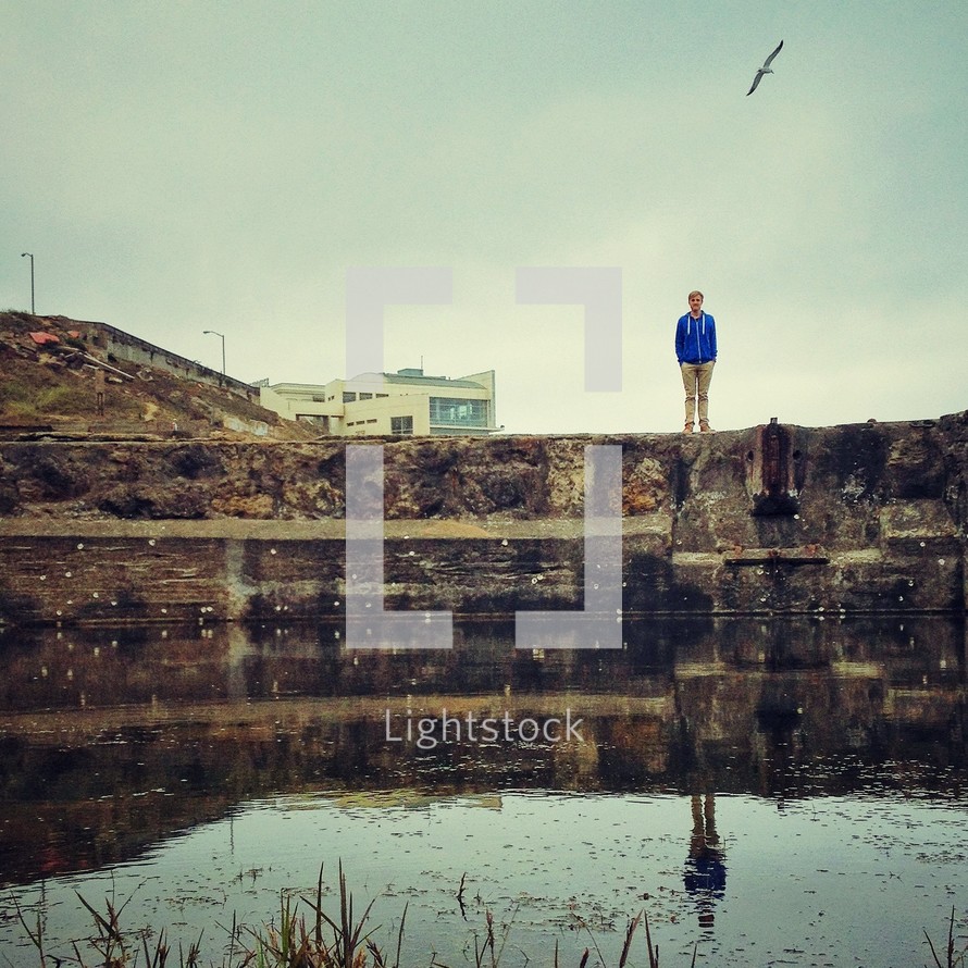 Man standing on rocks at edge of water with hill and building and flying bird in the background.