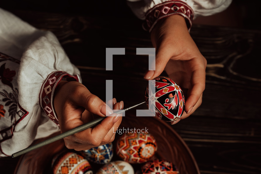 Ukrainian woman painting traditional ornamets on Easter egg - pysanka. Artist working in national costume. Preparation for Christian holiday. 