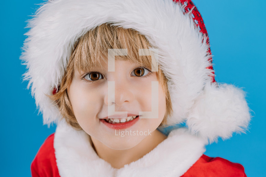Portrait of adorable boy in Santa Claus hat on blue studio background. Christmas celebration. Happy childhood, kid, lovely son. High quality photo
