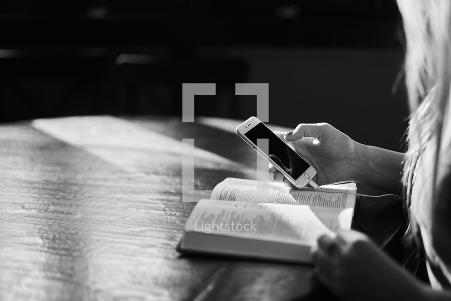 a woman looking at her cellphone while it is charging and an open Bible 
