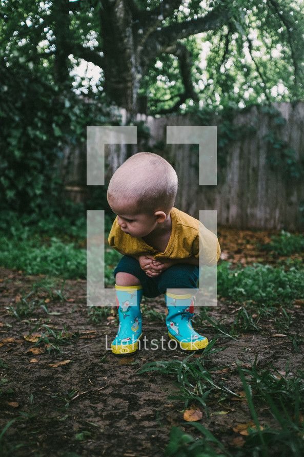 toddler boy in rain boots in a backyard playing in mud 