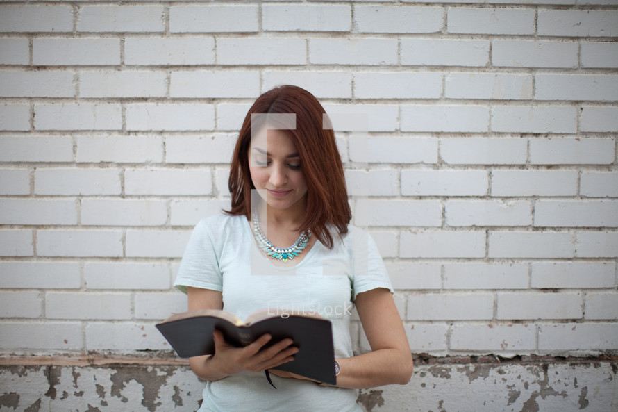 a young woman standing in front of a white wall and reading a Bible 