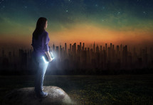 woman standing outdoors at night holding a glowing Bible looking out at a distant city 