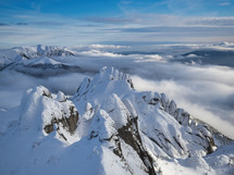 Aerial view of distant mountain peaks above clouds in clear sunny blue sky.