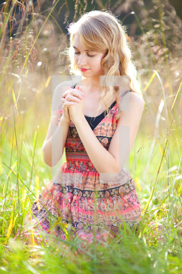 a blonde woman with praying hands kneeling in tall grass