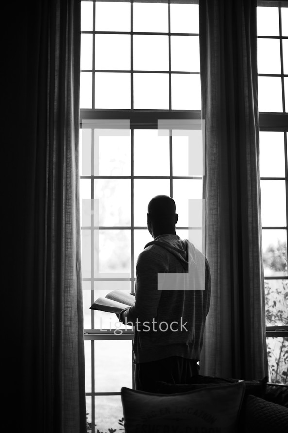 An African American man peers out a window with a Bible in his hands.