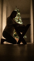 a woman kneeling to pickup a baby out of a manger in front of a Christmas tree 
