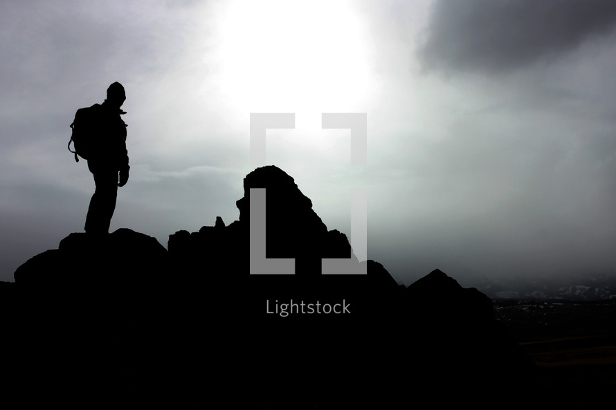 silhouette of a hiker standing on a rock under a cloudy sky