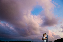 A bride and groom embrace outdoors under a cloudy sky.