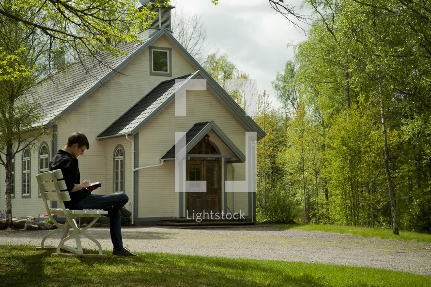 a man sitting on a bench in front of a chapel reading a Bible 