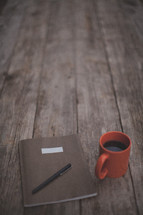 A pen and notebook next to an orange coffee mug on a wooden table