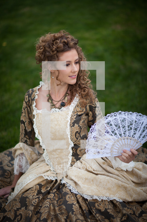 Woman in a Victorian dress with a lace fan sitting outside.