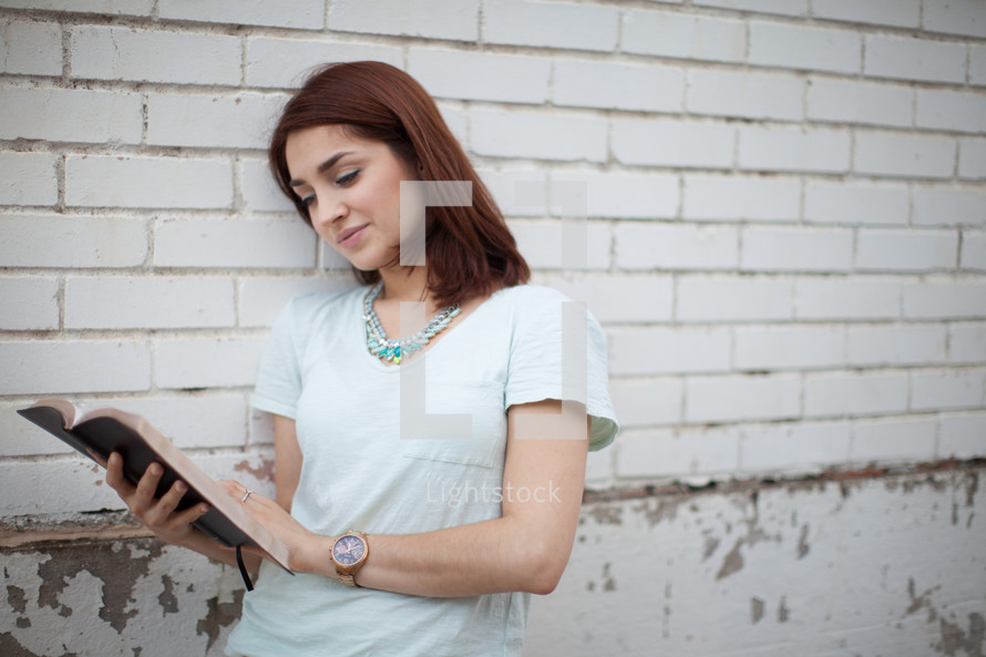 a young woman reading a Bible standing in front of a white wall 
