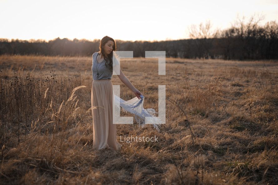 a woman with a long skirt and shaw standing in a field of dried grasses at sunset 
