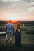 a couple holding hands on a hill with backs to the camera at sunset 