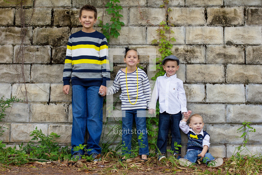 Four children holding hands and standing by a stone wall.