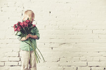 Boy hiding his face behind a bouquet of flowers standing in front of a brick wall.