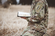 soldier kneeling in a field praying holding a Bible 