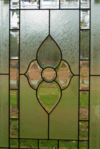 A cut glass window with stained glass pattern showing the outdoors and green grass peering through a cut glass window. 