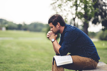 a man praying sitting outdoors with a Bible on his lap 