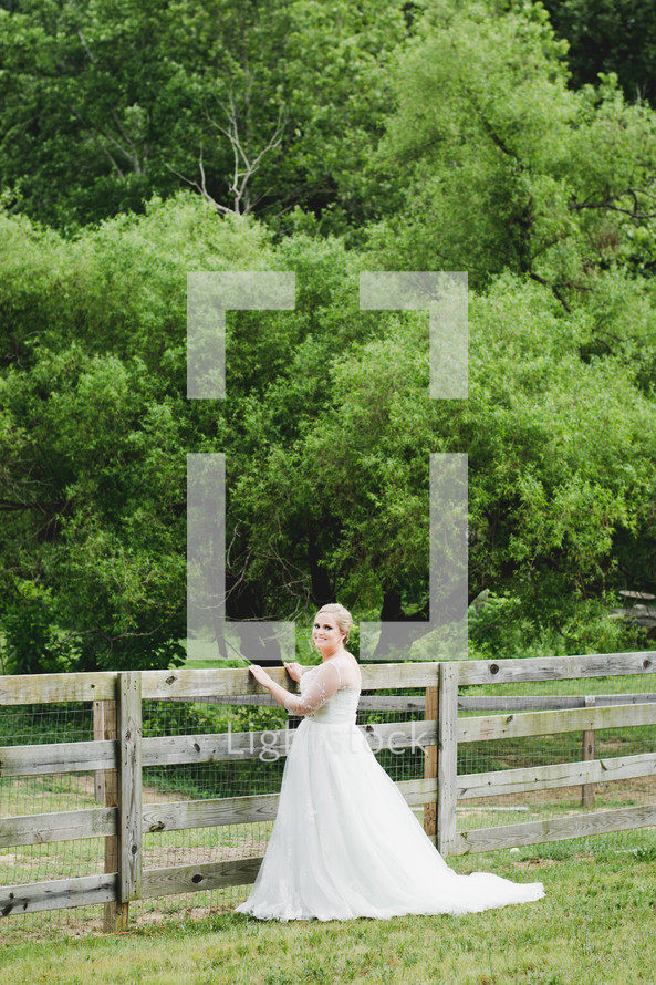 a bride standing in a field by a fence 