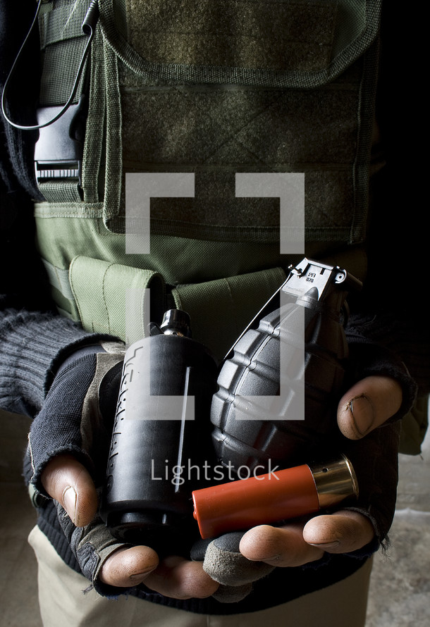 Man in military garb holding flashbang, fragmentation grenade, and shotgun cartridge.