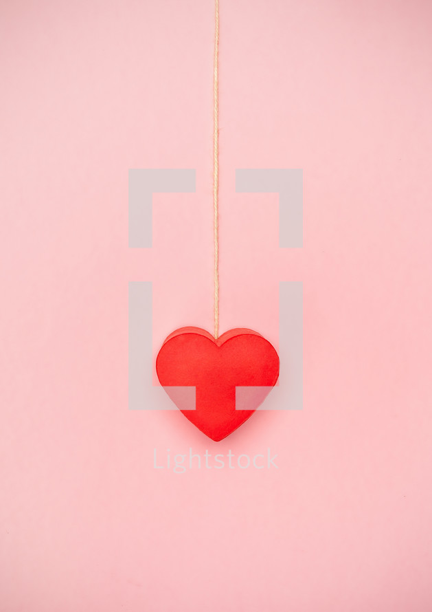 Heart hanging to a string of twine on pink background. Love concept hanging by a thread