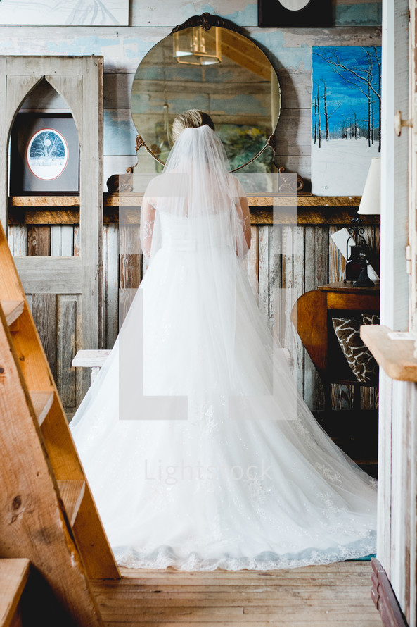 A bride standing in front of a round mirror in a rustic room.