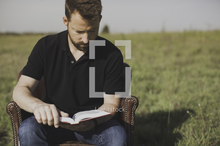 a man sitting in a chair outdoors reading a Bible 