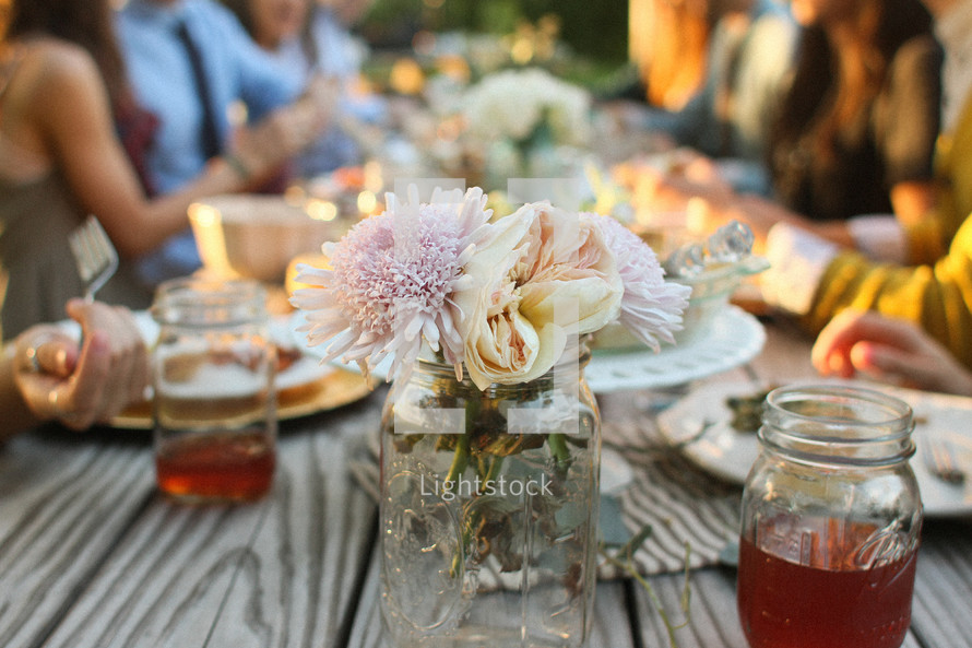 Friends sharing a meal together at a picnic table, with a mason jar of flowers and tea, outside.