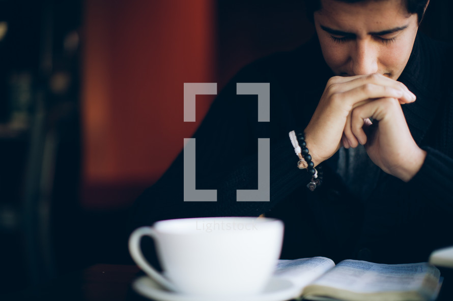 coffee mug and Bible resting on a table in front of a man in prayer 