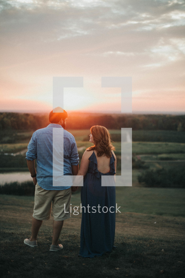 a couple holding hands on a hill with backs to the camera at sunset 