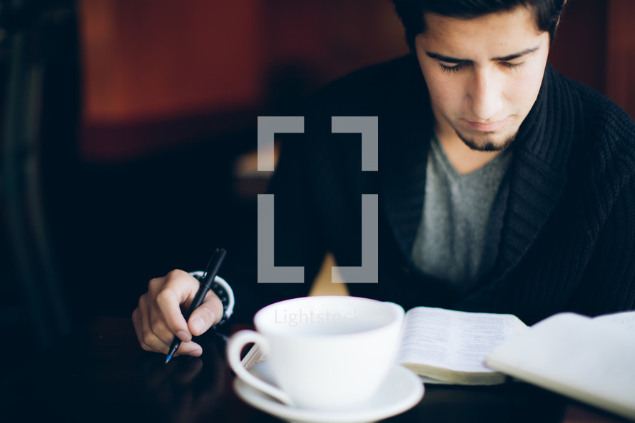 man reading a Bible and holding a pen sitting in a coffee house 