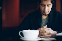 man with his hands held in prayer over a Bible and a coffee mug